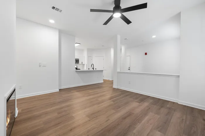 Modern open-concept living space featuring elegant hardwood floors, a sleek ceiling fan, and a cozy fireplace, leading to a stylish kitchen with white cabinetry.