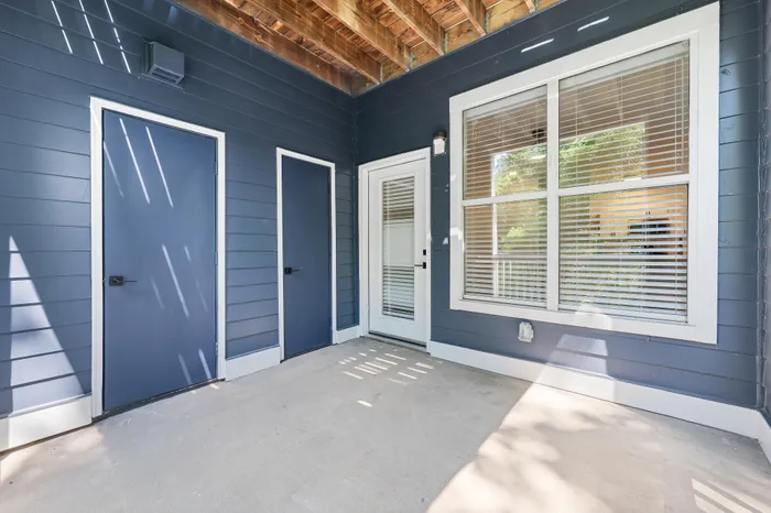 Charming outdoor patio space with modern blue paneling, dual doors, and large sunlit window with blinds.