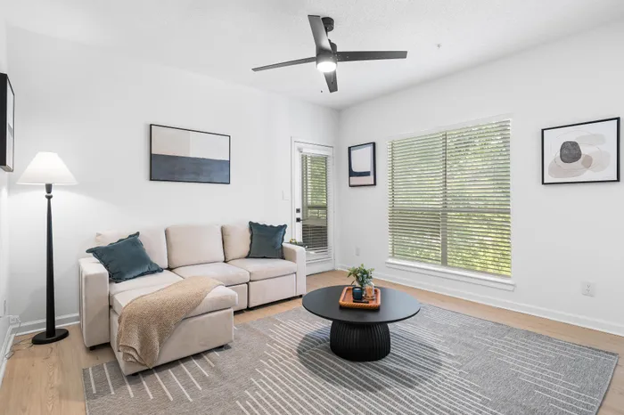 Bright and inviting living room featuring a cozy beige sectional sofa, sleek black coffee table, modern art, and large windows with lush greenery view.