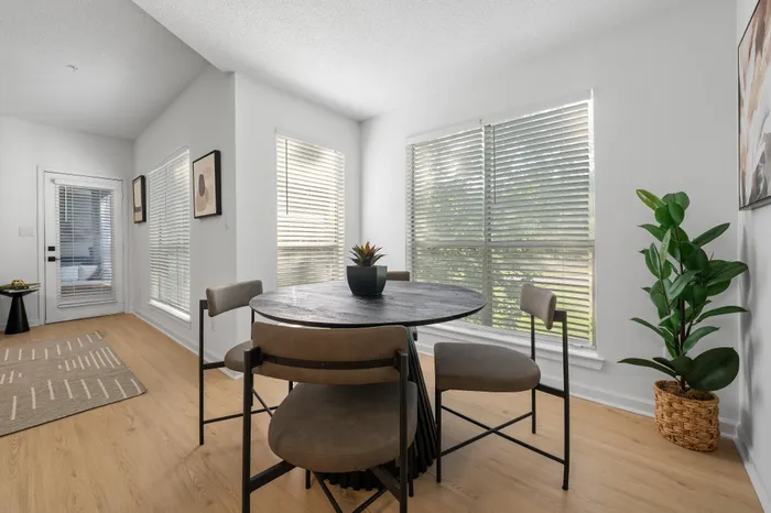 Bright dining area with a modern round table, stylish chairs, and large sunlit windows showcasing serene outdoor views.