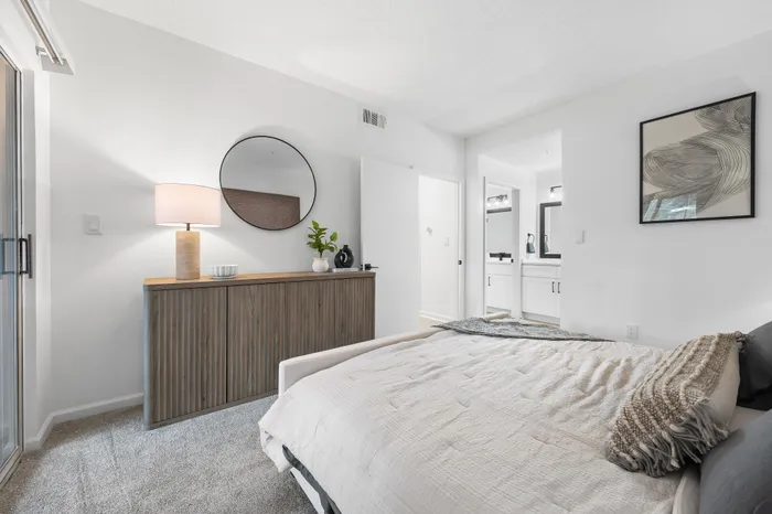 Bright and elegant bedroom with modern decor, featuring a stylish cabinet, round mirror, and cozy bed, bathed in natural light.