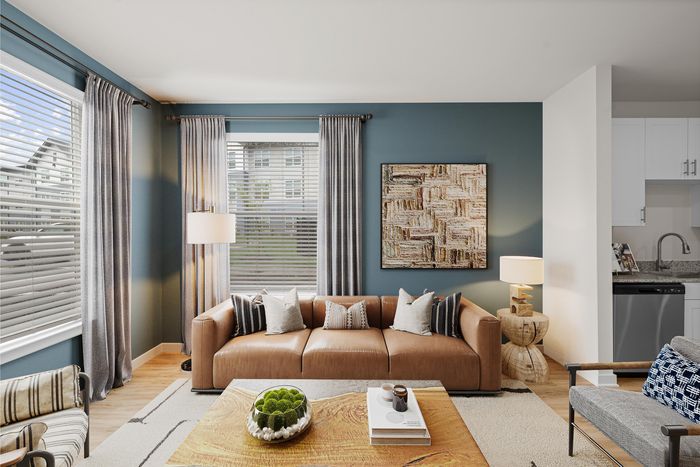 Modern living room with a leather sofa, chic artwork on a teal accent wall, large windows with elegant drapes, and a glimpse of a sleek kitchen.