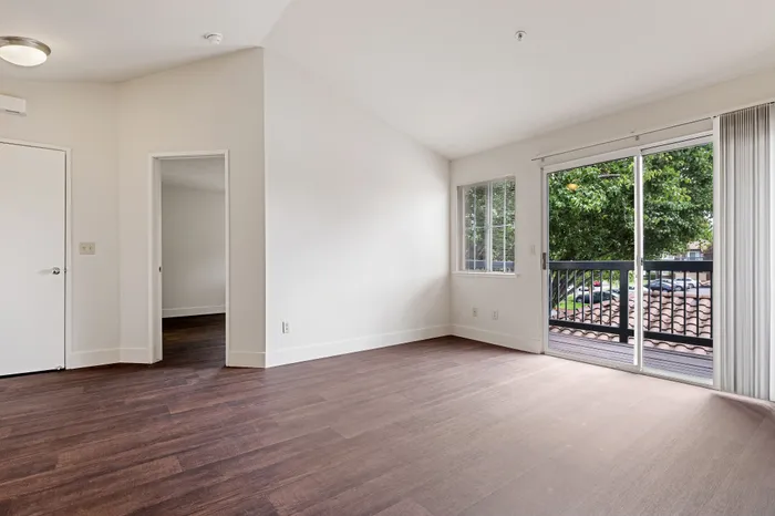 Bright and spacious room with rich wood flooring, white walls, and a large glass door leading to a charming balcony overlooking lush greenery.