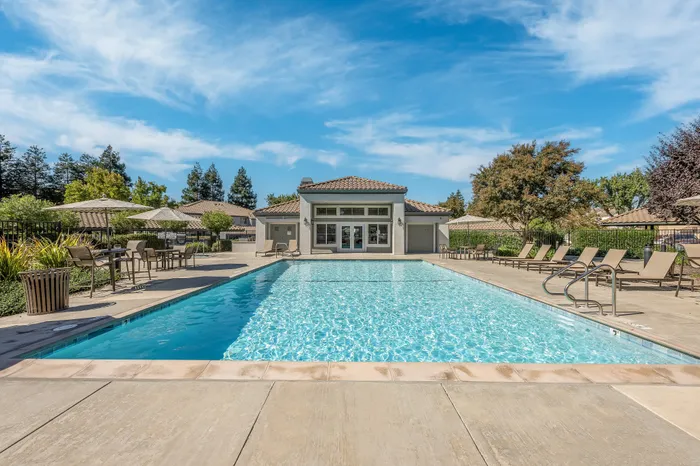 Luxurious outdoor pool area with stylish lounge chairs and shaded seating, surrounded by lush greenery and modern architecture against a bright blue sky.