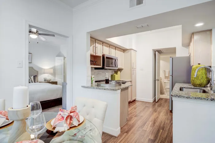 Elegant apartment interior showcasing an inviting kitchen with granite countertops, a cozy dining area set for two, and a glimpse of a modern bedroom.