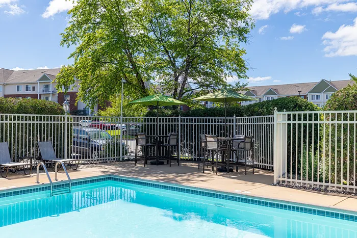 Inviting outdoor pool area with lounge chairs and shaded tables, surrounded by lush greenery and modern apartments.
