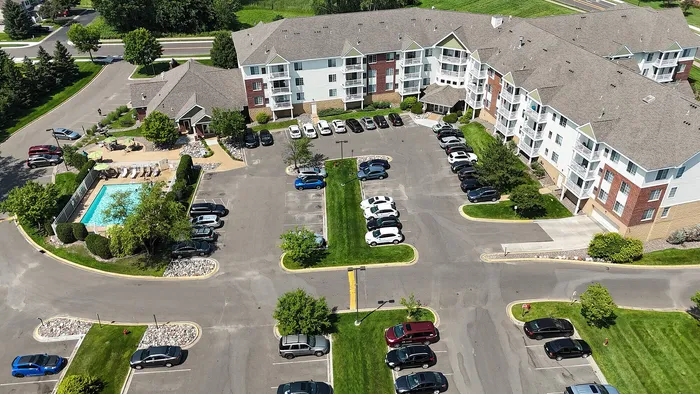 Aerial view of a modern apartment complex featuring lush green surroundings, a shimmering outdoor pool with lounge chairs, and spacious parking areas.
