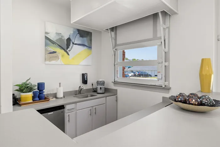 Modern kitchenette with sleek countertops, vibrant artwork, and natural light from a large window.