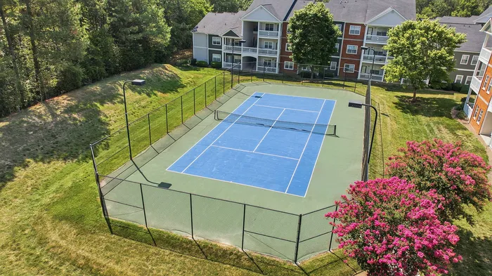 Charming apartment complex with a vibrant tennis court surrounded by lush greenery and blooming pink trees, offering a perfect blend of recreation and natural beauty.
