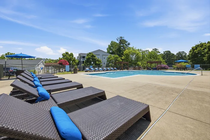 Scenic apartment community pool with cushioned loungers, surrounded by lush greenery and bright blue skies.