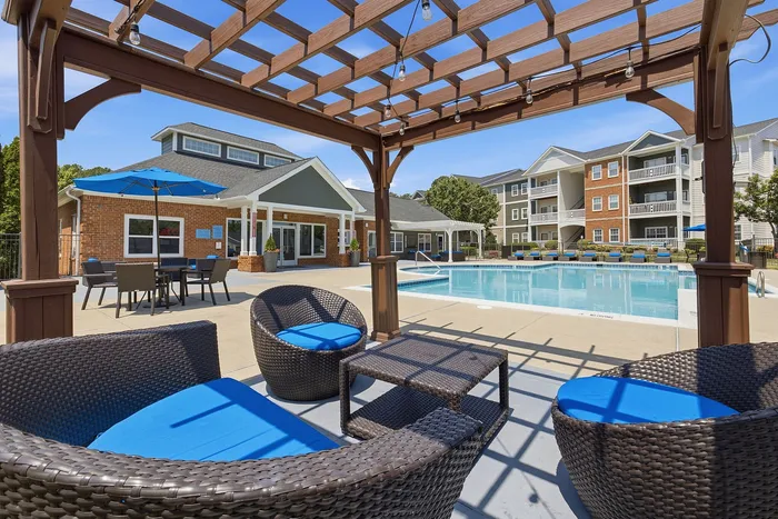Inviting outdoor lounge with wicker seating under a wooden pergola, overlooking a crystal-clear pool surrounded by elegant residential buildings.
