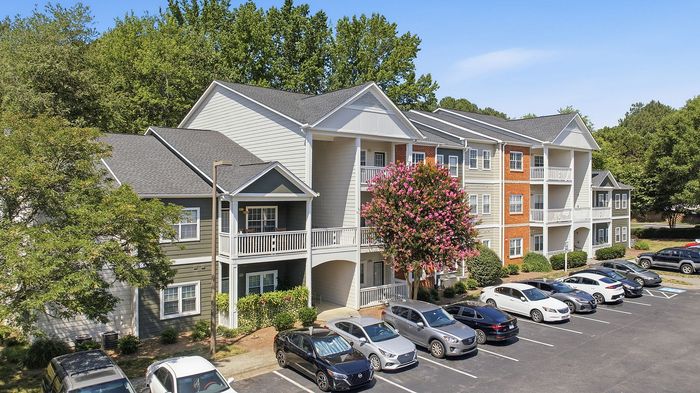 Charming three-story apartment complex with balconies, surrounded by lush greenery and vibrant flowering trees, featuring ample parking space.