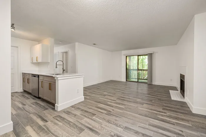 Modern open-concept living space featuring stylish wood flooring, sleek kitchen with elegant cabinetry, and a cozy fireplace next to a large glass door opening to a lush view.