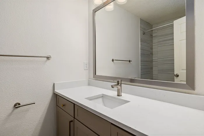 Modern bathroom featuring a sleek quartz countertop, elegant faucet, and a large framed mirror, complemented by stylish brushed nickel accessories and a striped tile shower.
