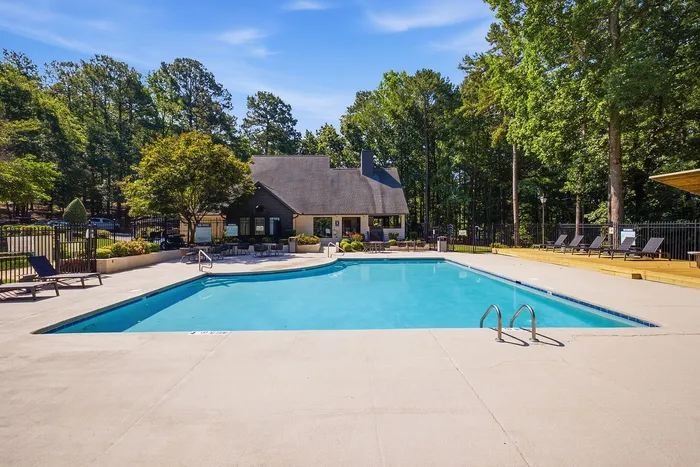 Beautiful swimming pool area with lush greenery, lounge chairs, and a charming clubhouse on a sunny day.