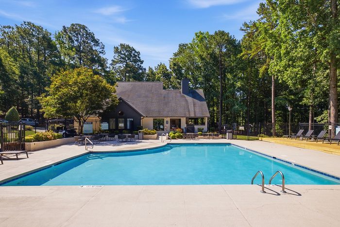 Inviting outdoor pool area surrounded by lush greenery, featuring comfortable lounge chairs and a charming clubhouse under a bright blue sky.