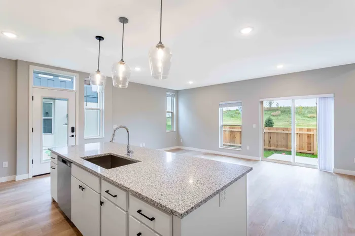 Bright, modern open-concept living space with a sleek granite kitchen island, elegant pendant lighting, and large windows offering a tranquil view of the lush green backyard.