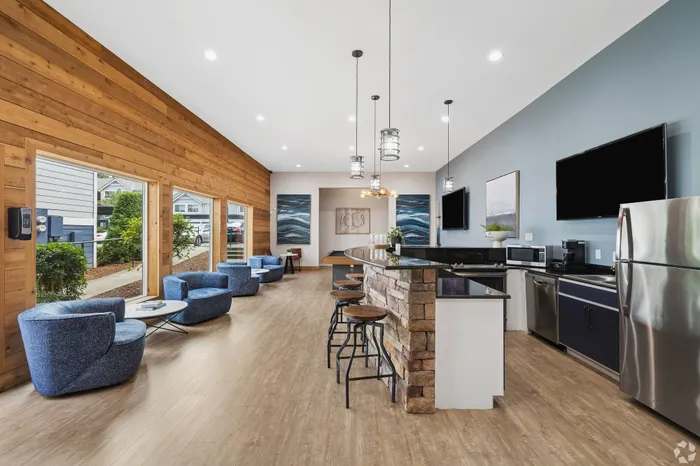 Modern community room with stylish stone bar, sleek kitchen appliances, blue accent chairs, and large windows offering natural light.