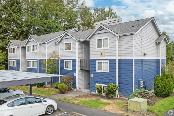 Charming two-story apartment building with modern blue and gray facade, surrounded by lush greenery and ample parking space.