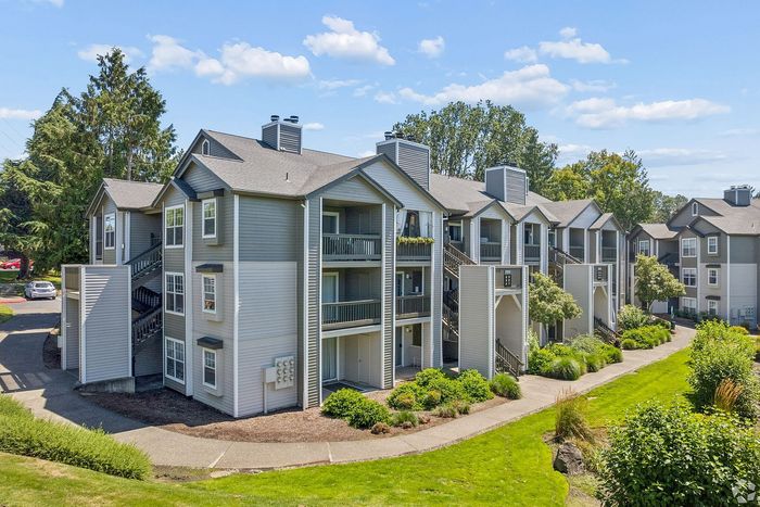 Charming multi-level apartment complex with modern gray and white exterior, surrounded by lush greenery and landscaped pathways.