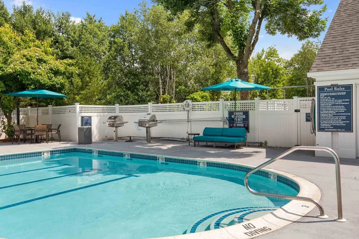 Charming outdoor pool area with inviting seating, shaded umbrellas, and modern barbecue grills, surrounded by lush greenery.