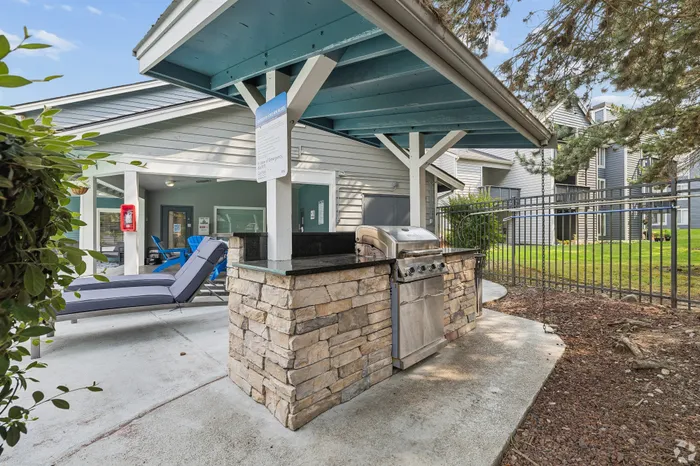 Charming outdoor area with a stylish stone BBQ grill, relaxing lounge chairs, and a cozy seating space under a light blue shaded patio.