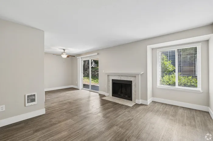 Bright living room with elegant wood flooring, modern ceiling fan, and a charming fireplace, featuring large windows and a patio door for abundant natural light.