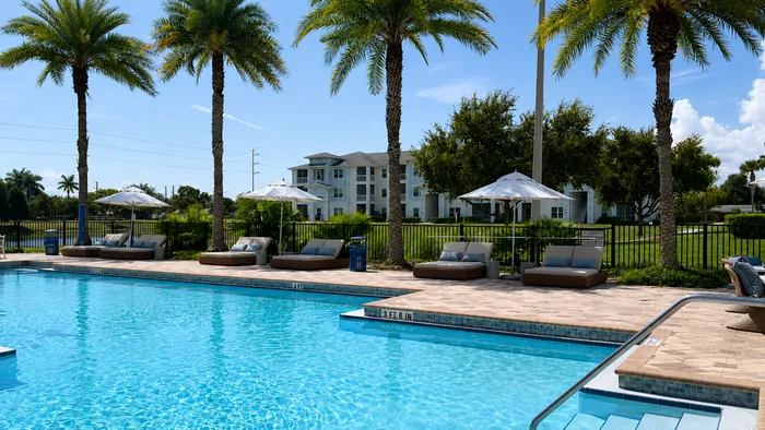 Luxurious apartment complex with a crystal-clear pool, elegant sun loungers, and swaying palm trees against a blue sky.