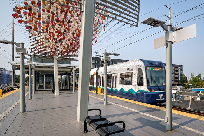 Modern urban light rail station with artistic canopy, featuring a sleek train arriving on a sunny day.
