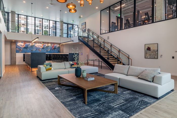 Modern lobby featuring stylish lounge area with plush sofas, elegant wood coffee table, artistic wall decor, and an impressive staircase leading to a glass-walled fitness center.