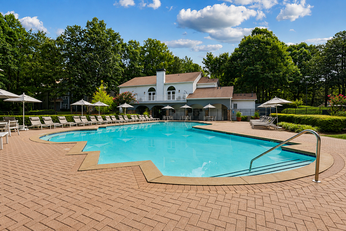 Inviting outdoor pool area with luxurious loungers and umbrellas, surrounded by lush greenery and a charming clubhouse under a sunny sky.