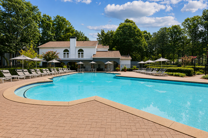 Luxurious outdoor pool area with elegant lounge chairs, surrounded by lush greenery and a charming clubhouse under a bright blue sky.