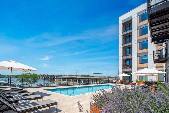 Luxurious apartment community with a stunning outdoor pool area overlooking a serene waterfront, complemented by elegant lounging spaces and vibrant landscaping under a clear blue sky.