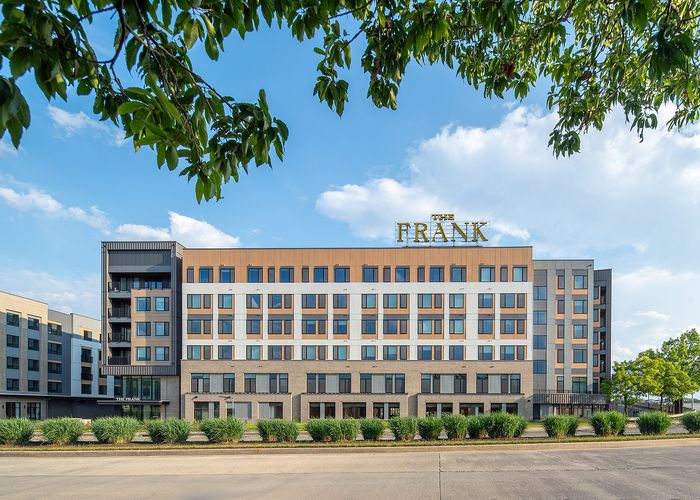 Modern mid-rise building 'The Frank' under a clear blue sky, surrounded by lush greenery and showcasing sleek architectural design.