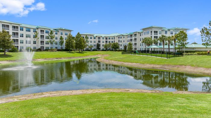 Serene apartment complex with elegant architecture, set amid lush greenery and a picturesque pond with a fountain.