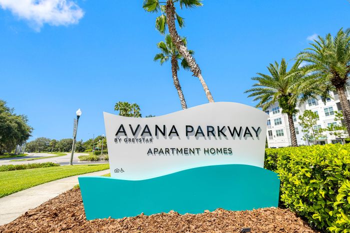 Inviting sign for Avana Parkway Apartment Homes set amidst lush greenery and palm trees under a bright blue sky.