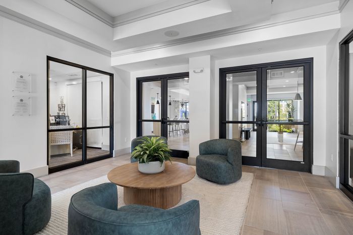 Chic lobby area featuring modern armchairs and a stylish center table with a potted plant, complemented by sleek glass doors in a welcoming apartment complex.