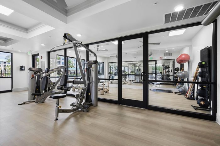 Modern gym space with state-of-the-art fitness equipment, bright natural lighting, and sleek glass partition separating workout zones.