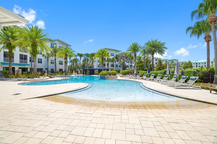 Luxurious apartment complex featuring a sparkling resort-style pool surrounded by palm trees and comfortable lounge chairs, set against a bright blue sky.