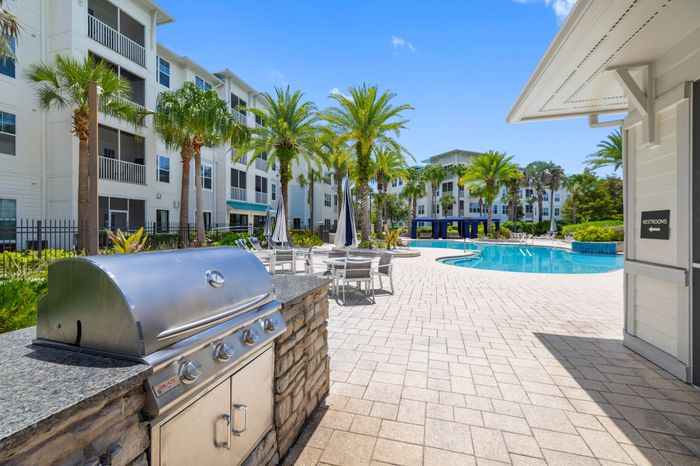 Luxurious apartment complex with a sparkling pool, palm trees, and outdoor barbecue area under clear blue skies.