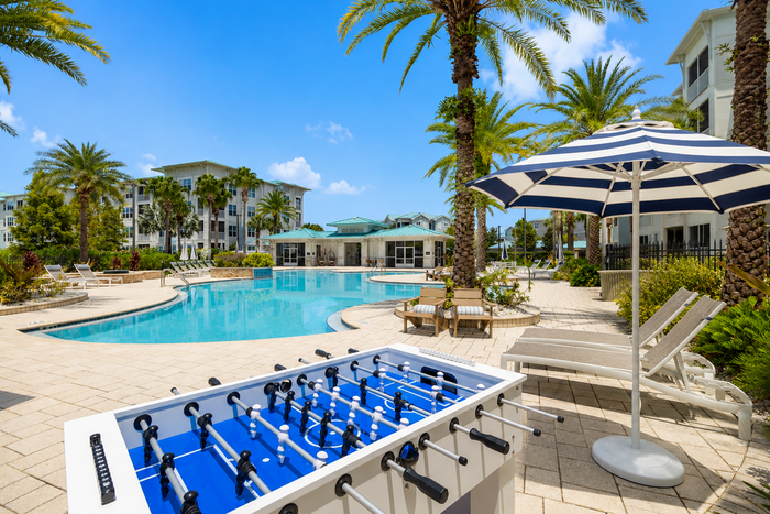 Luxurious resort-style apartment complex featuring a sparkling swimming pool, elegant sun loungers, and vibrant palm trees under a clear blue sky.