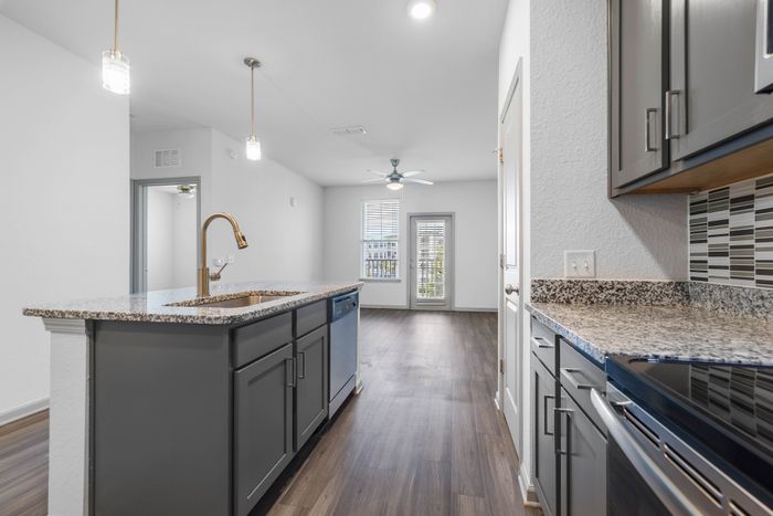 Chic apartment kitchen with granite countertops, modern appliances, and pendant lighting leading to a bright living area.