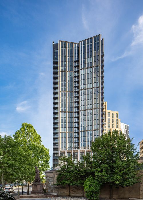 Stunning modern skyscraper with sleek glass facade, surrounded by lush green trees and cityscape under a bright blue sky.