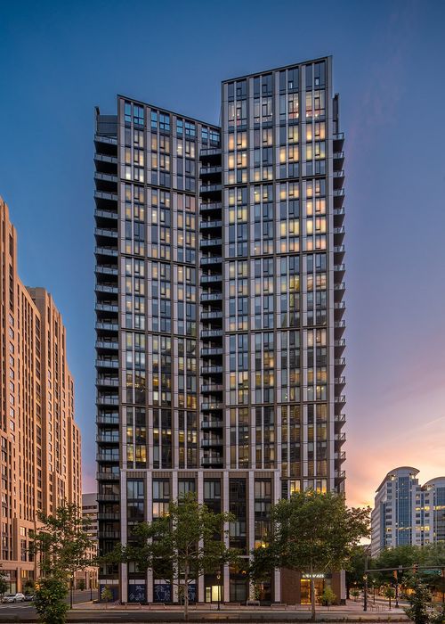 Modern high-rise apartment building with sleek glass fa&ccedil;ade, illuminated windows at dusk, surrounded by lush greenery and cityscape.