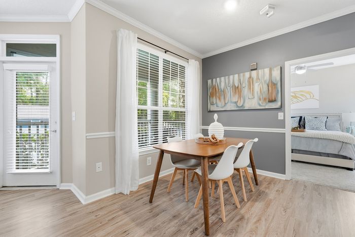 Bright dining area with a chic wooden table and modern chairs, featuring large windows and stylish wall art, seamlessly connecting to a cozy bedroom.