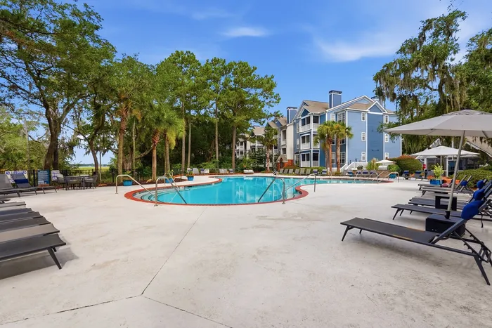 Inviting outdoor pool area surrounded by lush greenery and modern blue apartments, complete with lounge chairs and umbrellas for a relaxing retreat.