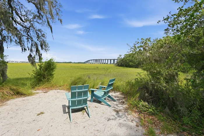 Scenic view of two teal chairs on a sandy spot overlooking lush green marshland with a bridge in the distance under a clear blue sky.
