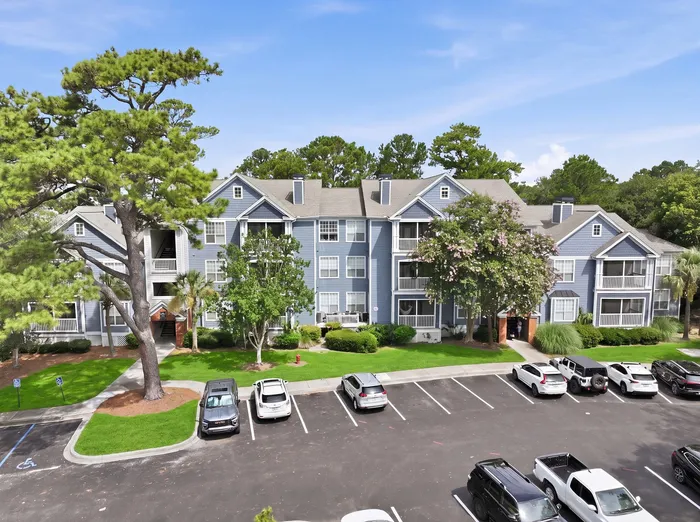 Charming blue apartment building with lush greenery and ample parking space, nestled in a serene setting under a clear blue sky.