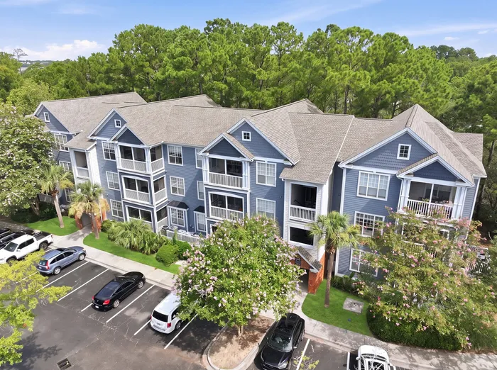 Charming modern apartment building with blue siding, lush landscaping, ample parking, and surrounded by vibrant green trees.