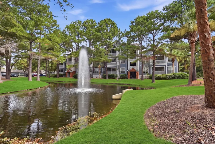 Charming apartment complex surrounded by lush greenery and featuring a serene pond with a central fountain.